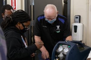 A masked white male man instructs a masked Black female student at an HVAC unit inside the Tidewater Tech campus