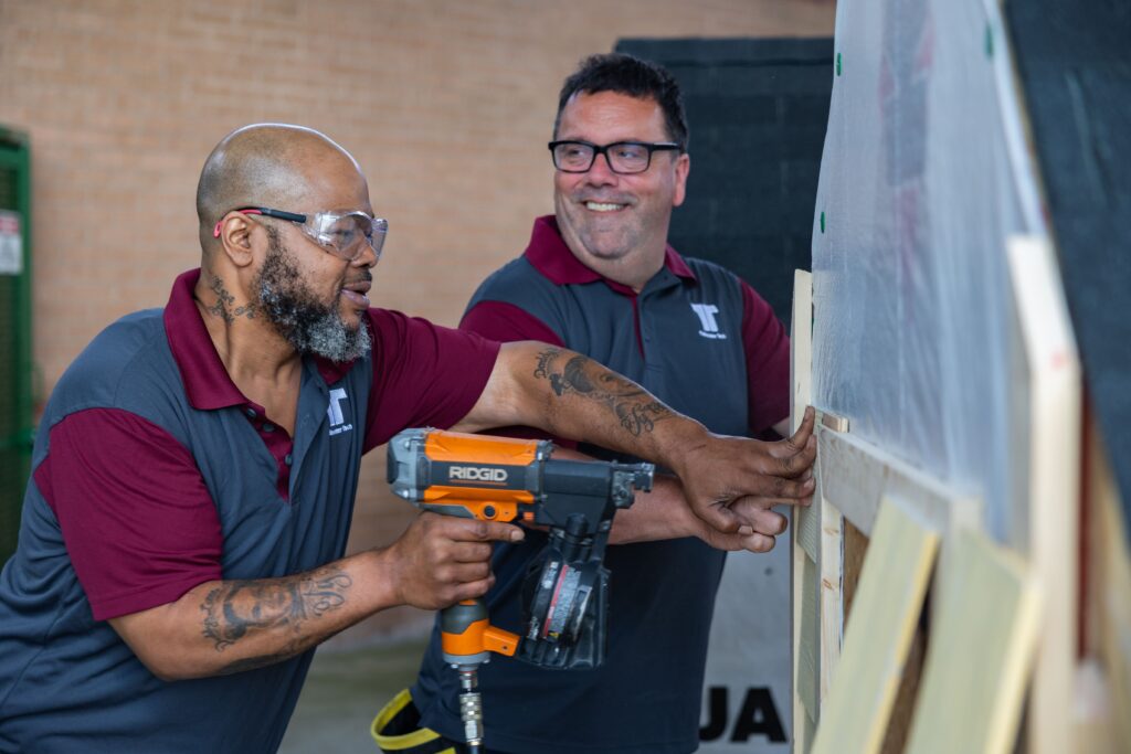 Two Tidewater Tech students working together during a construction training program, using a nail gun on a wooden frame as part of hands-on learning.