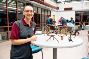 Woman HVAC student at Tidewater Tech with copper pipe sculptures, highlighting women in skilled trades.